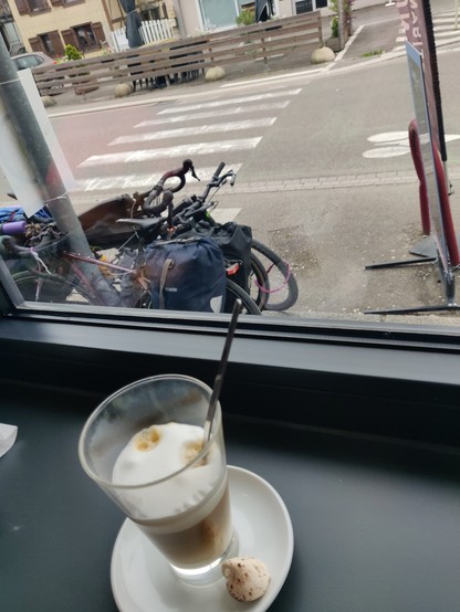 Ein Latte Macchiato im Vordergrund. Perspektive leicht von oben. Blick durchs Café-Fenster. Draußen stehen die bepackten Reisräder.