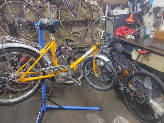Ein Klapprad in einem Montageständer. Daneben steht längs zu einer Werkbank ein Rennrad. Im Hintergrund sind mehrere alte Stahlfahrräder.