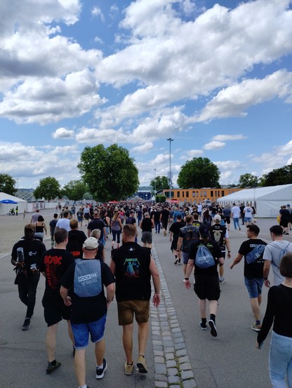 Ein Menschenmenge, überwiegend in schwarzen T-Shirts und kurzen Hosen laufen Richtung einer Bühne die ganz weit hinten steht uns kaum zu erkennen ist. Links und rechts sind weiße Zelte zu sehen.