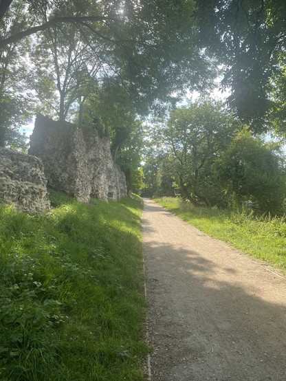 Foto eines Schotterwegs, eingesäumt von Wiese und Bäumen. Auf der linken Seite des Weges ist eine Reihe alter Säulen, welche aus Steinen und einer Art Zement aufgebaut sind. Sie sind verwittert, aber gepflegt