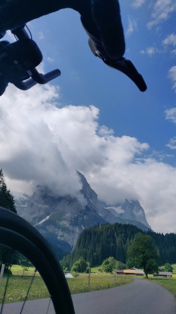 Aus der Perspektive meines rechten Knies Blick zwischen Vorderrad und Lenker auf die (gerade flache) kleine Straße durch ein alpines Hochtal. Hohe Berge in Wolken im Hintergrund. Sommerliche Stimmung.