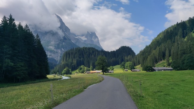 Die kleine Straße schlängelt sich vor mir durch ein alpines Hochtal. Links Wald und Bach. Hohe Berge in Wolken im Hintergrund. Sommerliche Stimmung.