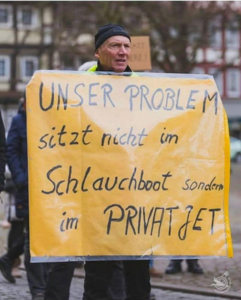 Demonstrant, der folgendes Schild trägt:

UNSER PROBLEM
sitzt nicht im
Schlauchboot sondern
im PRIVATJET